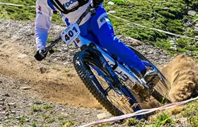 Un ciclista de montaña, vestido con un maillot azul y blanco, con casco y el dorsal 401, desciende por un sendero de tierra rodeado de hierba y terreno rocoso.