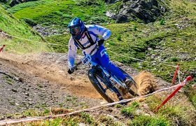 Un ciclista de montaña vestido de azul y blanco baja por un sendero de tierra, levantando polvo, con colinas cubiertas de hierba y rocas al fondo.