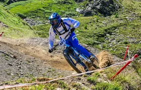Un ciclista con equipación azul y blanca baja en bicicleta de montaña por un sendero rocoso y de tierra en la ladera cubierta de hierba, levantando una nube de polvo a su paso.