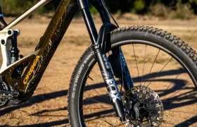 Primer plano de la rueda delantera y la horquilla de suspensión de una potente bicicleta de montaña con motor Bosch Performance Line CX, captada en un camino de tierra con árboles de fondo.