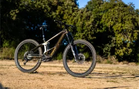 Una moderna bicicleta de montaña con suspensión total y un potente motor Bosch Performance Line CX se encuentra en un camino de tierra, rodeada de hierba y árboles bajo un cielo azul despejado.