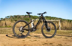 Una potente bicicleta eléctrica Bosch Performance Line CX en negro y plata se encuentra en un campo de tierra con una portería y hierba alta al fondo bajo un cielo azul claro.