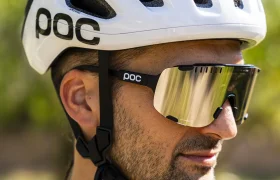 Hombre al aire libre con un casco de ciclismo polivalente blanco de la marca POC y unas gafas de sol grandes reflectantes, vestido con un maillot de ciclismo ligero adecuado para el ciclismo de carretera o gravel, con vegetación difuminada al fondo.