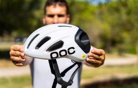 Una persona sostiene al aire libre un casco de ciclismo polivalente POC de color blanco, con una correa negra y una pegatina amarilla de MIPS, con árboles al fondo; perfecto tanto para salidas por carretera como por grava.