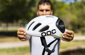 Una persona sostiene al aire libre un casco de ciclismo POC blanco con rejillas de ventilación negras y una correa ajustable, con árboles y césped al fondo; ideal como casco polivalente tanto para aventuras en grava como en carretera.