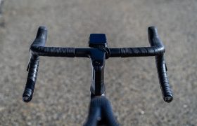 Primer plano del manillar negro de una bicicleta, envuelto con cinta Silca Nastro Cuscino para mayor comodidad y un tacto superior, con un ciclocomputador instalado, fotografiado al aire libre sobre una superficie pavimentada.