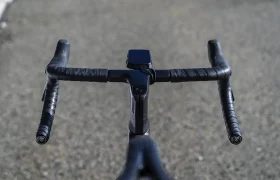 Primer plano del manillar de una bicicleta negra envuelto con cinta Silca Nastro Cuscino para mayor comodidad y tacto, con un ciclocomputador montado en el centro, en una carretera asfaltada y desierta.