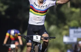Un ciclista vestido con un maillot blanco con rayas arco iris, que recuerda a la leyenda del MTB Nino Schurter, levanta ambos brazos en señal de victoria mientras cruza la línea de meta, con otro ciclista al fondo.