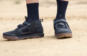Una persona está de pie sobre un suelo arenoso y lleva zapatillas negras Leatt ProClip 4.0 de ciclismo con cierre de esfera y correa y calcetines negros.