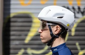 Persona con casco POC Cytal blanco y gafas transparentes, mirando hacia la izquierda. El vibrante fondo de grafitis difuminados le da un toque urbano, mientras que el casco de carretera garantiza seguridad y estilo.