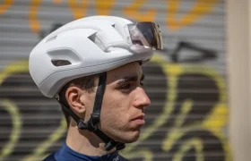 Una persona que viste un casco de carretera blanco POC Cytal, con un par de gafas de sol encima, se para con confianza frente a una pared cubierta de grafitis.