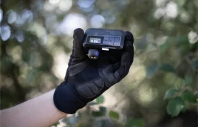 Una mano enguantada sostiene un dispositivo digital compacto Trek Air Rush con pantalla de visualización, al aire libre con un follaje verde borroso de fondo, perfecto para inflado de ruedas sobre la marcha.