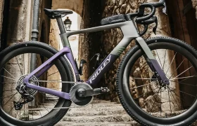 Una elegante bicicleta de carretera, la Ridley Kanzo Fast 2.0, con un llamativo cuadro morado y gris, está aparcada en una escalera de adoquines entre antiguos edificios de piedra. Su diseño aerodinámico destaca en medio del paisaje histórico, lista para deslizarse por cualquier ruta con estilo y velocidad.