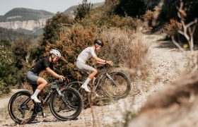 Dos ciclistas, equipados con casco y ropa de ciclismo, circulan en bicicletas de montaña Ridley Kanzo Fast 2.0 con neumáticos de 58 mm cuesta arriba por un sendero rocoso rodeado de matorrales y colinas.