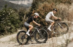 Dos ciclistas, con casco y ropa deportiva, suben en sus bicicletas de montaña Ridley Kanzo Fast 2.0 por un sendero de grava rodeado de árboles y colinas, afrontando con seguridad el terreno gracias a sus cuadros aerodinámicos y a los neumáticos de 58 mm, que les proporcionan mayor estabilidad.