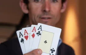 Un hombre sostiene cuatro cartas, tres ases de diferentes palos y una carta en blanco, con las cartas mirando hacia la cámara, en un evento de Alpinum TotalEnergies.