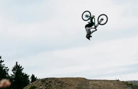 Robin Goomes realiza un backflip sobre una bicicleta de montaña encima de una rampa de tierra, luciendo gafas Ora y un casco Coron Air, con árboles y un cielo nublado de fondo.