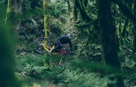 Un ciclista con ropa oscura realiza un truco Air-Over-Air en una MTB roja y amarilla, equipada con un RockShox Reverb AXS, en medio de un bosque denso y cubierto de musgo.