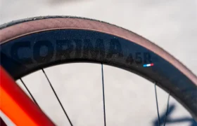Primer plano de una rueda de bicicleta en la que destaca la marca Ruedas Corima 45R en la llanta negra, famosa por su ligereza y rendimiento, con parte de un cuadro de bicicleta naranja visible al fondo.