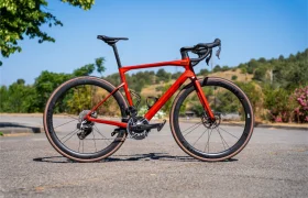Una bicicleta de carretera roja con Ruedas Corima 45R, manillar rebajado y frenos de disco está aparcada en una superficie pavimentada al aire libre, ofreciendo una ligereza y un rendimiento impresionantes con árboles y colinas de fondo.
