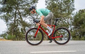 Un ciclista con casco y ropa de ciclismo conduce una bicicleta de carretera roja con Ruedas Corima 45R por una carretera asfaltada, disfrutando de la ligereza y el rendimiento entre los árboles del fondo.