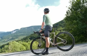 Un ciclista con casco y ropa de ciclismo junto a una bicicleta de carretera equipada con Corima Essentia 42 ruedas endurance, mirando hacia unas colinas verdes y boscosas bajo un cielo parcialmente nublado.