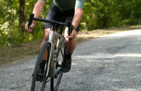 Un ciclista con casco y gafas de sol monta en una bicicleta de carretera equipada con ruedas Corima Essentia 42 por un camino pavimentado a través de una zona verde y arbolada.