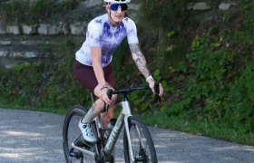 Un ciclista con casco blanco, gafas de sol y maillot morado conduce una bicicleta equipada con ruedas Corima Essentia 42 por una carretera asfaltada junto a un muro de piedra y una frondosa vegetación.