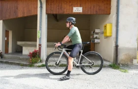 Un ciclista con casco y maillot verde permanece de pie junto a una moderna bicicleta de carretera equipada con Corima Essentia 42 ruedas endurance frente a un edificio rústico con un cartel en el que se lee "Place du Bascil".