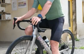 Un ciclista con casco, gafas de sol y ropa de ciclismo se sienta en una bicicleta de carretera equipada con gravilla Corima Essentia 42 ruedas frente a un edificio con marcos de ventanas amarillos.