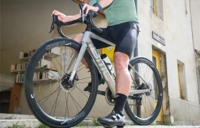 Una persona vestida de ciclista, a horcajadas sobre una bicicleta de carretera con ruedas Corima Essentia 42, perfectas para recorridos de grava y resistencia, frente a un edificio con una estantería y ventanas de marcos amarillos.