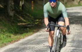 Un ciclista con casco y gafas de sol conduce una bicicleta de carretera equipada con ruedas carretera cuesta arriba por un camino rural sombreado y rodeado de árboles.