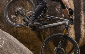Un ciclista, vestido con ropa negra y casco, baja en bicicleta de montaña por una pendiente rocosa y empinada, manteniendo el equilibrio con la rueda delantera levantada.
