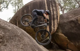 Un ciclista de montaña, con casco, sube por una empinada pared rocosa entre grandes cantos rodados en una zona boscosa.