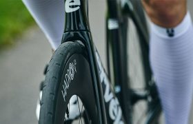 Primer plano de una rueda de bicicleta negra HADRON³ Ultimate 650 con el texto "HANDSON", parte de una bicicleta Cervélo, y un ciclista con calcetines blancos a rayas en un camino pavimentado, diseñado para la aerodinámica máxima dentro del límite UCI.
