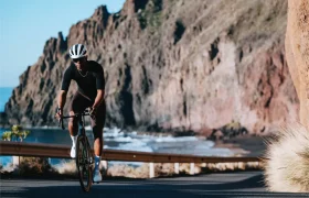 Un ciclista con casco y ropa de ciclismo negra conduce una bicicleta de ruta Zipp 2026 por una carretera costera con acantilados y el océano de fondo, buscando rodar más rápido en esta ruta escénica.