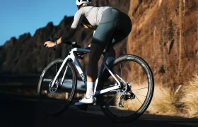 Un ciclista circula en una bicicleta de carretera blanca con ruedas Zipp 2026 por una carretera asfaltada junto a acantilados rocosos y hierba seca, usando casco y equipo de ciclismo para rodar más rápido.