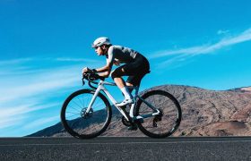 Un ciclista con casco y equipo de ciclismo recorre una bicicleta de ruta Zipp 2026 por una carretera asfaltada, disfrutando de una conectividad inteligente con montañas y un cielo azul de fondo.