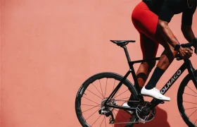 Una persona que viste pantalones cortos de ciclismo de color rojo monta una bicicleta de carretera Colnago negra con ruedas Zipp 2026, lista para rodar más rápido contra una pared sólida de color melocotón.
