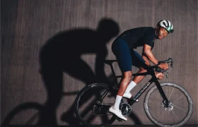 Un ciclista vestido con traje y casco negros conduce una bicicleta de ruta Zipp 2026 contra un fondo oscuro y liso, proyectando una sombra prominente en la pared, listo para rodar más rápido.