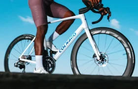 Una persona vestida con ropa de ciclista monta una bicicleta de carretera Colnago blanca con ruedas Zipp 404 S bajo un cielo azul claro; la imagen, tomada desde un ángulo bajo, destaca la búsqueda de la velocidad.