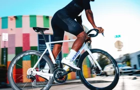Un ciclista vestido con ropa deportiva negra pedalea en una bicicleta de carretera Colnago blanca equipada con ruedas Zipp 404 S, pasando a toda velocidad junto a un edificio de colores en un día soleado.