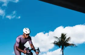 Un ciclista, vestido con un traje morado y un casco blanco, pedalea en una bicicleta de carretera equipada con ruedas Zipp 404 S al aire libre, bajo un cielo azul parcialmente nublado, con una palmera al fondo, haciendo gala tanto de velocidad como de estilo.