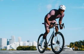 Un ciclista, equipado con casco y ropa de ciclismo, pedalea al aire libre en una bicicleta de carretera Zipp 404 S, disfrutando de la velocidad con el perfil urbano y un cielo azul y despejado como telón de fondo.