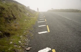 En una carretera con niebla cerca de Conor Pass, una persona anda en bicicleta a través de la niebla, flanqueada por un terreno cubierto de hierba y marcas viales visibles.
