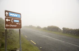 Carretera con niebla y un cartel que dice "An Chonair", que muestra las direcciones y las distancias a través del Paso Conor. Una vegetación escasa y muros de piedra bordean los costados de esta ruta panorámica.