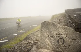 Un ciclista recorre una carretera con niebla cerca de Conor Pass y pasa junto a una piedra con la inscripción "Sistersip Damgean Ui Chúise Dingle Daingean Uí Chúis".