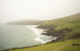 Paisaje costero brumoso con colinas verdes y acantilados que se unen al mar en Conor Pass. Las olas rompen suavemente contra la costa rocosa, mientras que a lo lejos se ven algunos edificios rurales.