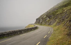 La sinuosa carretera costera, que recuerda al pintoresco Conor Pass, está bordeada por un muro de piedra y acantilados cubiertos de hierba bajo un cielo brumoso.