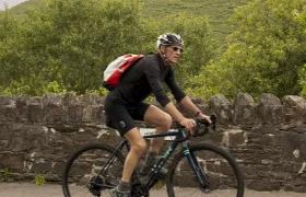 Un ciclista con casco y mochila recorre la ruta panorámica cerca de Conor Pass, pedaleando junto a un muro de piedra con una colina cubierta de hierba al fondo.